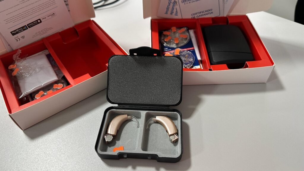 A imagem mostra duas caixas contendo aparelhos auditivos. Ambas as caixas são brancas com detalhes vermelhos e contêm os aparelhos auditivos em um estojo preto com revestimento interno de espuma. Além dos aparelhos, as caixas incluem: * *Manual de instruções:* Um pequeno livreto com instruções de uso e informações sobre o aparelho auditivo. A imagem mostra partes do manual em português, indicando instruções de uso e um certificado de garantia. * *Baterias:* Pequenas baterias para os aparelhos auditivos, provavelmente em embalagens individuais. A imagem mostra várias baterias em embalagens laranja. * *Acessórios:* Outros acessórios, como protetores de ouvido ou outros componentes pequenos, também estão incluídos. * *Estojo de transporte:* Um estojo compacto e protetor para armazenar os aparelhos auditivos quando não estão em uso. Este estojo é preto e tem compartimentos para cada aparelho. Em resumo, as imagens mostram dois novos aparelhos auditivos completos com todos os acessórios e materiais de informação necessários para o seu uso. A presença de dois conjuntos sugere que podem ser um par para uso em ambos os ouvidos.