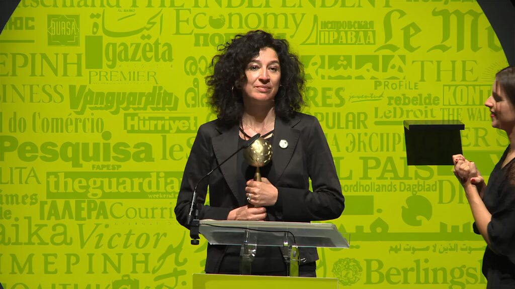  Mulher de cabelos cacheados e escuros, vestida de preto, segura um troféu dourado em forma de globo, enquanto discursa em um púlpito com microfone. Ela está em um palco iluminado por fundo amarelo com nomes de diversos jornais e revistas impressos. Ao lado direito, uma mulher com vestido preto observa a cena.