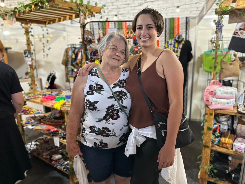 A imagem mostra duas mulheres sorrindo e abraçadas em um ambiente de feira, cercadas por bancas de artesanato colorido. Elas parecem felizes e descontraídas, aproveitando o momento juntas. Ao fundo, há bolsas, bijuterias e itens decorativos expostos. O cenário transmite um clima acolhedor e festivo.