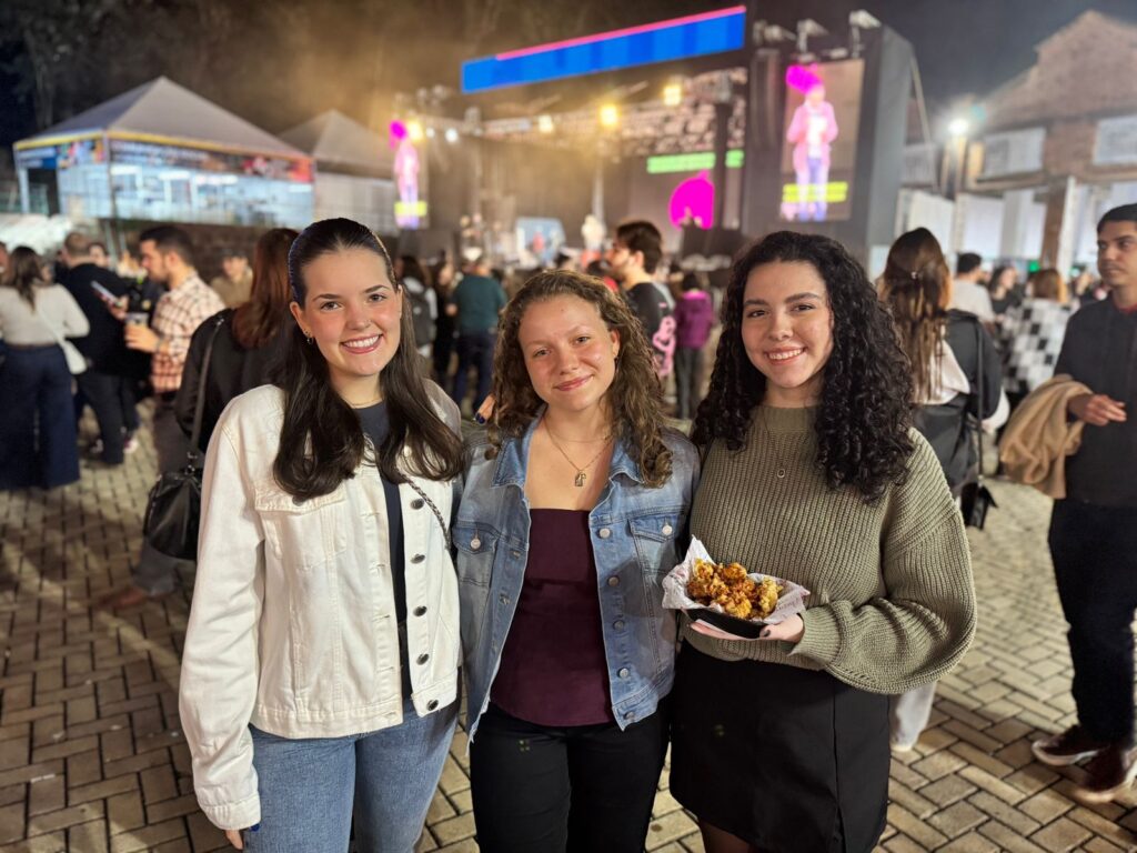 A imagem mostra três pessoas reunidas em um evento ao ar livre, possivelmente um festival. No fundo, há um palco iluminado com uma grande tela exibindo um artista, e a área parece cheia de espectadores. Também há tendas e outras estruturas visíveis. Um dos indivíduos está segurando um prato de comida, o que sugere um ambiente descontraído e social. O clima parece festivo e animado, com muita gente aproveitando o momento.