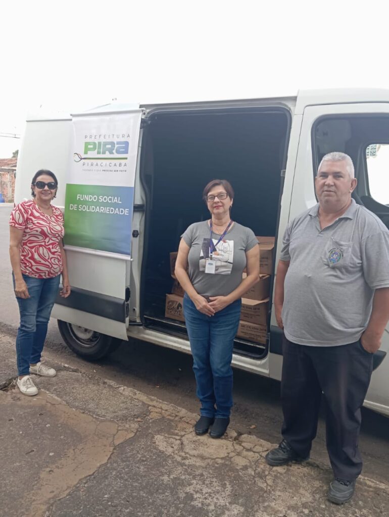 A imagem mostra três pessoas em pé na frente de uma van branca. A van tem um letreiro na lateral que diz "PREFEITURA PIRA PIRACICABA FUNDO SOCIAL DE SOLIDARIEDADE". Duas mulheres e um homem estão em pé na frente da van. As mulheres estão usando jeans e blusas, e o homem está usando uma camisa cinza e calças pretas. Parece que eles estão distribuindo suprimentos ou fazendo trabalho de caridade. 