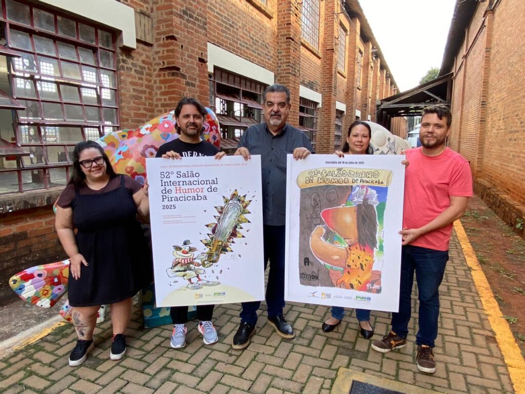 Um grupo de cinco pessoas, quatro homens e uma mulher, posa em frente a um prédio de tijolos aparentes segurando dois cartazes. O da esquerda divulga o 52º Salão Internacional de Humor de Piracicaba, com ilustração de um palhaço serrando uma serra gigante com flores coloridas. O da direita, o cartaz do Salãozinho de Humor, apresenta uma figura infantil estilizada com traços coloridos. Todos sorriem e estão posicionados em um corredor externo entre os prédios do Engenho Central.