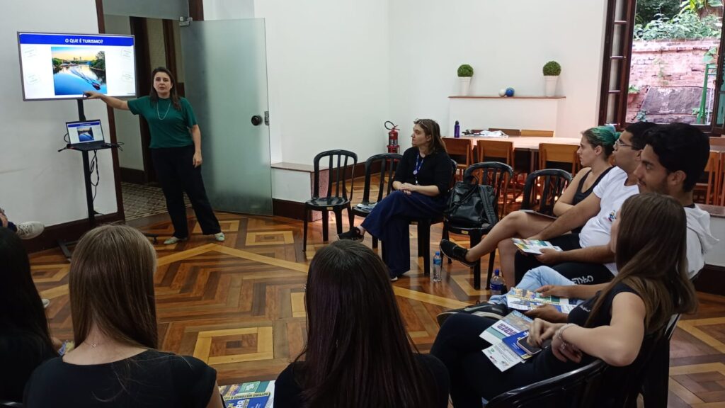 Uma mulher está ministrando uma palestra para um grupo de pessoas sentadas em círculo.
A composição da cena mostra um grupo de pessoas em um espaço amplo, provavelmente uma sala de aula ou um local de reunião. As pessoas estão sentadas em cadeiras de plástico pretas ao redor de uma área central. Uma mulher de blusa verde está em pé, demonstrando algo em um monitor de computador. O grupo de participantes está em uma posição atenta e envolvida na apresentação, demonstrando interesse e escuta. A cena parece ser em um momento de aprendizado, onde a atenção dos participantes está focalizada na mulher que fala.As figuras principais são a mulher que está dando a apresentação e um grupo de pessoas. A mulher que fala está usando uma blusa verde e calças pretas, está em pé no centro, com um tom de voz normal, como se fosse uma conversa. Os participantes parecem atentos e interessados no conteúdo da apresentação. A maioria dos participantes são adultos jovens, e suas expressões denotam interesse e concentração na palestra.O estilo da imagem é fotográfico, representando um evento real, sem efeitos artísticos. O foco está na interação entre os participantes, mostrando uma dinâmica de aprendizagem.O local parece ser um espaço interno, com chão de madeira escura e paredes brancas. Há uma janela em um dos cantos, mostrando uma visão do lado de fora, mas o foco principal está nos participantes e na apresentação em andamento. A iluminação é uniforme, e não há sombras muito fortes, ideal para facilitar a visão dos participantes. A atmosfera é de concentração e aprendizagem, como em uma sala de aula.