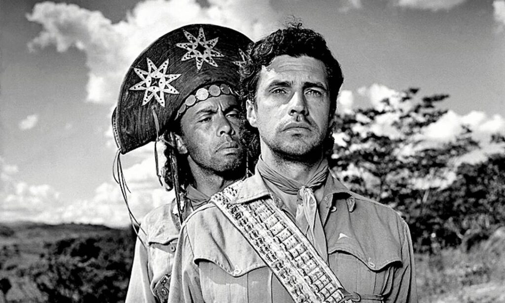 Imagem em preto e branco do filme O Cangaceiro, dirigido por Lima Barreto. Em primeiro plano, dois homens estão de pé olhando para a frente. O homem da frente usa traje típico de cangaceiro com cartucheira e lenço no pescoço, enquanto o homem ao fundo usa chapéu de couro adornado com estrelas. Ao fundo, vegetação típica do sertão e céu com nuvens.