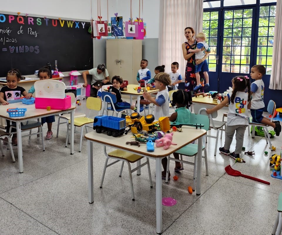 A imagem mostra uma sala de aula de pré-escola vibrante e ativa. Crianças pequenas estão sentadas em mesas pequenas, envolvidas em atividades de jogo com brinquedos como caminhões de brinquedo e animais de plástico. Algumas crianças estão sentadas em suas mesas, enquanto outras estão interagindo umas com as outras. Há duas professoras na sala, uma interagindo com as crianças e outra observando. O quadro-negro na parede de trás exibe o alfabeto e algumas palavras escritas em português, sugerindo uma escola brasileira. A sala tem uma atmosfera alegre e acolhedora, com paredes coloridas e uma iluminação natural abundante.