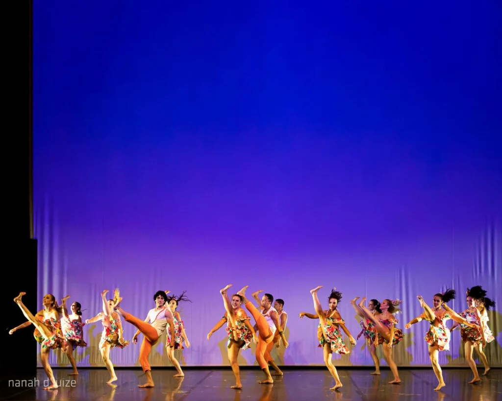 Fotografia colorida de um espetáculo de dança com cerca de 20 bailarinos e bailarinas no palco. O fundo é um grande painel iluminado em tons degradê de azul e lilás. Os dançarinos usam figurinos coloridos e vibrantes, com saias floridas e blusas claras. Estão em movimento, com pernas erguidas e braços expressivos, compondo uma coreografia dinâmica. A cena transmite energia, leveza e sincronismo. No canto inferior esquerdo lê-se o crédito "nanah duize".