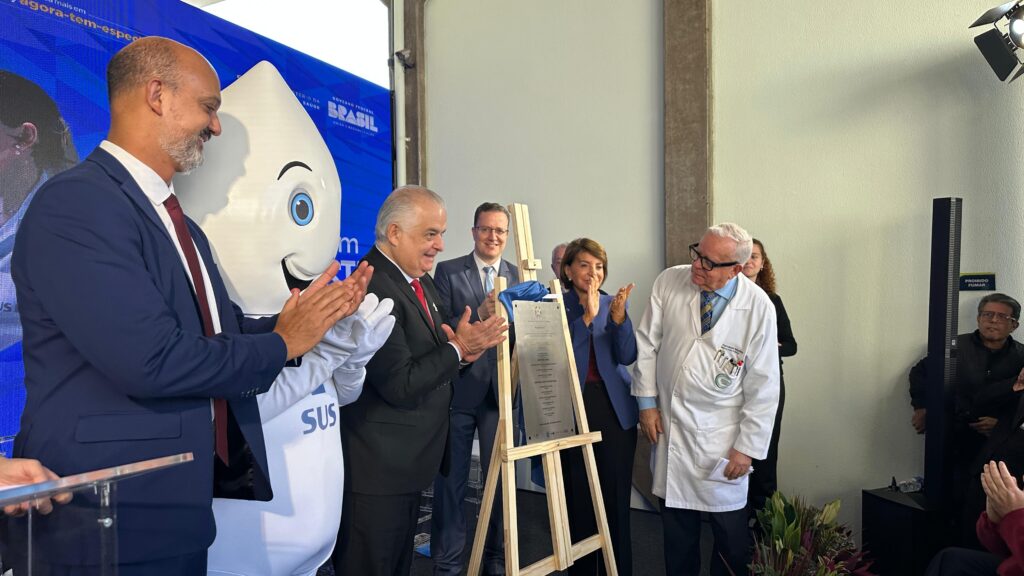 A imagem mostra um homem de terno falando em um púlpito. Ele está usando óculos e um microfone está posicionado na frente dele. Ao fundo, há uma mulher interpretando em Libras (Língua Brasileira de Sinais), uma mulher em uma roupa médica e outras pessoas sentadas. Um banner com a frase "Agora tem especialistas" é visível ao fundo. A cena parece ser uma conferência de imprensa ou um evento público, possivelmente relacionado à saúde, dada a presença da mulher em uniforme médico e a frase no banner. O homem de terno é provavelmente um orador ou palestrante, e a interprete de Libras garante a acessibilidade para pessoas surdas. As outras pessoas sentadas podem ser autoridades, jornalistas ou membros da audiência. A imagem sugere a divulgação de um programa ou iniciativa governamental relacionada à disponibilização de especialistas médicos, dado o contexto visual e a frase no banner. A imagem é bem iluminada e nítida, permitindo uma boa visualização dos detalhes.