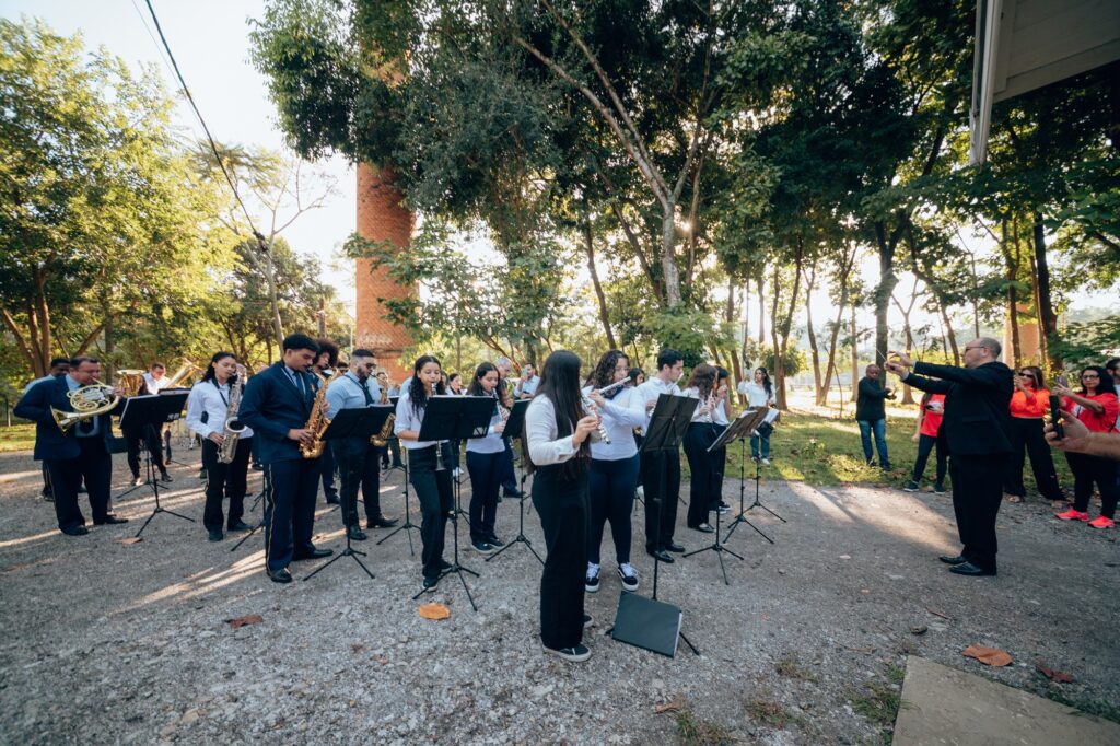 A imagem mostra um grupo de músicos se apresentando ao ar livre. Eles estão tocando diversos instrumentos, como saxofones, flautas e trompas, e estão organizados em um semicírculo, lendo partituras em suportes musicais. À direita, um regente conduz a performance. O cenário é uma área semelhante a um parque, com árvores e vegetação ao fundo. Algumas pessoas estão de pé assistindo à apresentação.