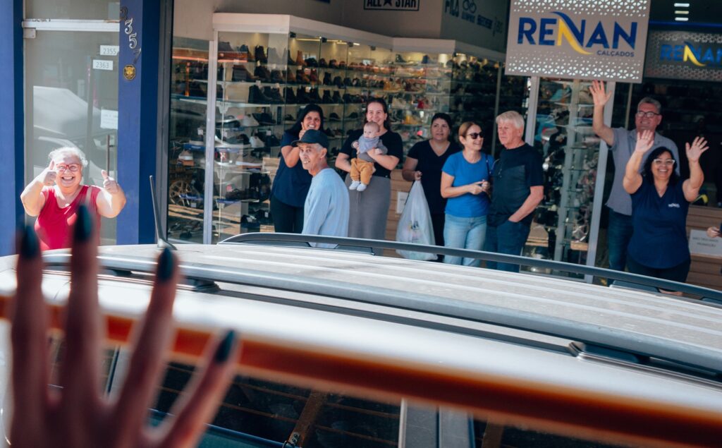 A imagem mostra um grupo de pessoas em frente a uma loja chamada "RENAN Calçados". A fachada da loja tem vidro e exibe diversos sapatos. As pessoas estão acenando e parecem estar cumprimentando alguém. No primeiro plano, há uma mão com unhas pintadas, sugerindo que a perspectiva é de dentro de um veículo. Os números "2355" e "2363" estão visíveis na moldura azul da porta da loja.