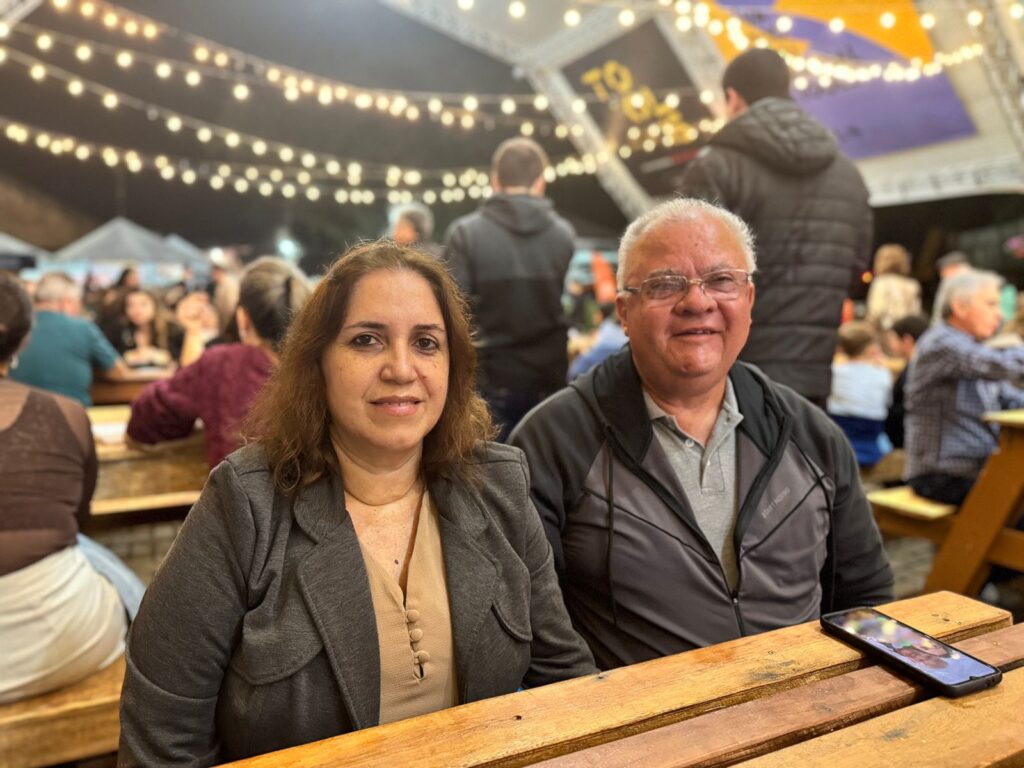 A imagem mostra duas pessoas sentadas em uma mesa de madeira em um ambiente que parece ser um evento ou festival ao ar livre. O fundo está iluminado com luzes suspensas e há vários participantes, alguns também sentados em mesas. Há um celular colocado sobre a mesa à frente das duas pessoas. O ambiente parece animado e social, com muitas pessoas reunidas e conversando.