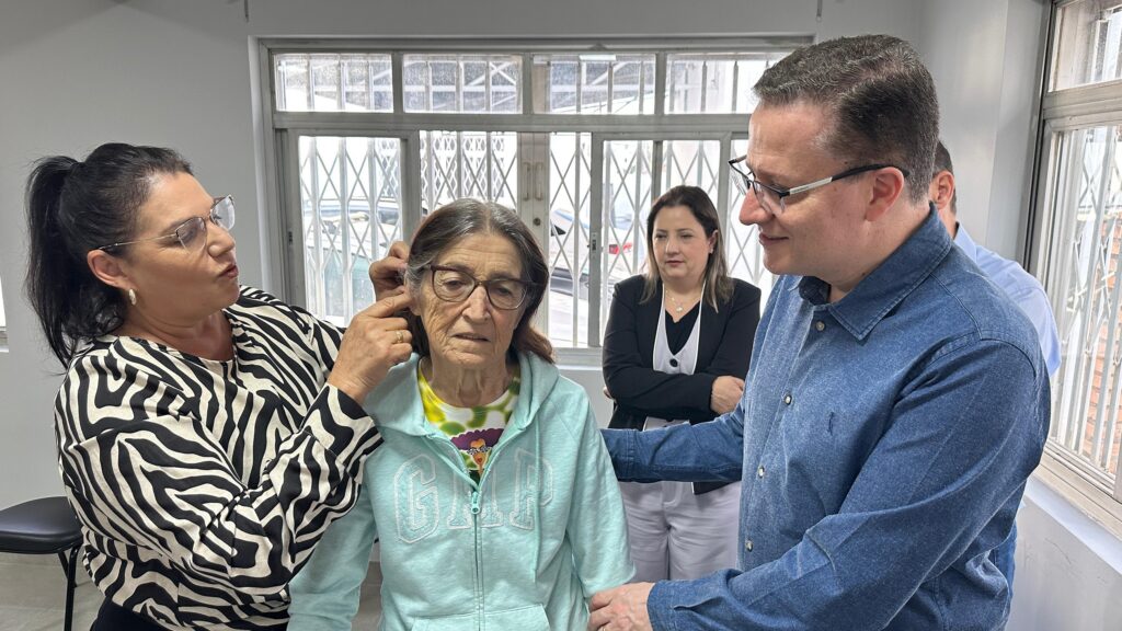 A imagem mostra uma cena em um ambiente que parece ser um consultório médico ou uma clínica. Há quatro adultos e uma senhora idosa no centro da imagem. Duas mulheres estão ajudando a senhora idosa, aparentemente colocando ou ajustando um aparelho auditivo. Uma das mulheres está vestida com uma blusa de zebra e a outra parece ser uma profissional de saúde ou assistente. Um homem, que parece ser um médico ou profissional de saúde, está envolvido no processo, ajudando a senhora. Uma outra mulher está observando a cena ao fundo. A cena sugere um atendimento de saúde auditiva, possivelmente a colocação ou verificação de aparelhos auditivos em uma paciente idosa. O ambiente é simples, com uma janela grande mostrando uma vista externa parcialmente visível. A iluminação é suave e natural, com uma atmosfera calma e profissional.