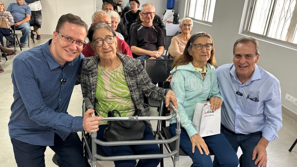 A imagem mostra um grupo de pessoas idosas sentadas em cadeiras em uma sala, possivelmente em um evento ou encontro. Há dois homens mais jovens sentados ao lado das senhoras idosas, um deles com o braço ao redor de uma das senhoras. As senhoras aparentam estar confortáveis e sorrindo para a câmera. Um dos homens parece estar ajudando uma das senhoras com uma bengala ou andador. O ambiente parece ser um centro comunitário, clínica ou local de atendimento a idosos. No fundo, outras pessoas idosas estão sentadas em cadeiras, algumas com expressões mais sérias. A imagem sugere um ambiente de cuidado e apoio para a terceira idade. Há uma bolsa de papel com uma marca visível na imagem, possivelmente indicando um tipo de assistência ou patrocínio ao evento. A imagem transmite uma sensação de comunidade e cuidado com os idosos.