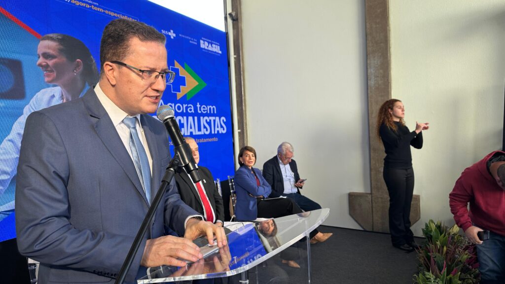 A imagem mostra uma coletiva de imprensa ou evento semelhante, com um homem falando ao microfone em um palco. Há um banner atrás dele que diz "Agora tem ESPECIALISTAS", indicando que o evento provavelmente está relacionado a um anúncio sobre a disponibilidade de especialistas médicos. Há outras pessoas na foto, incluindo outras autoridades ou profissionais de saúde, sentadas atrás dele. O banner também menciona um site do governo brasileiro (gov.br/saude). Uma mulher está de pé à direita, parecendo auxiliar na organização do evento. A imagem sugere um anúncio governamental de melhorias no acesso a especialistas médicos no Sistema Único de Saúde (SUS) do Brasil. O homem falando provavelmente é um oficial do governo, possivelmente um ministro da saúde ou um alto funcionário, fazendo o anúncio oficial. A presença de outras pessoas no palco indica que o evento é formal e importante. A imagem, por si só, não fornece detalhes sobre os tipos específicos de especialistas, o escopo geográfico do anúncio ou outras informações relevantes.