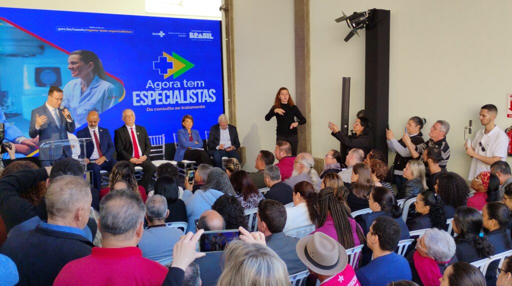A imagem mostra um evento público, provavelmente uma conferência de imprensa ou um anúncio governamental, focado na área da saúde. Há várias pistas que apontam para isso: * *Banner Promocional:* Um grande banner azul e amarelo com a frase "Agora tem ESPECIALISTAS" e o logotipo do Ministério da Saúde do Brasil é claramente visível. A frase "Da consulta ao tratamento" indica o foco no acesso a especialistas médicos. Também há um endereço web (gov.br/saude/agora-tem-especialistas), sugerindo uma iniciativa governamental. * *Palestrantes:* Várias pessoas estão sentadas em uma mesa de palco, aparentemente como palestrantes ou autoridades. Um homem está usando um microfone, sugerindo um discurso ou apresentação formal. Pelo menos uma mulher está usando um uniforme ou roupa que sugere uma posição na área de saúde. * *Público:* Uma grande audiência está sentada em cadeiras brancas, prestando atenção aos palestrantes. Algumas pessoas estão usando celulares para tirar fotos ou gravar o evento. * *Interpretação em Libras:* Uma mulher está usando linguagem de sinais (Libras), indicando que o evento está sendo acessível a pessoas com deficiência auditiva. * *Contexto:* A presença de um extintor de incêndio e equipamentos de som e iluminação sugere um local preparado para eventos públicos. Em resumo, a imagem retrata um evento oficial do governo brasileiro, provavelmente relacionado a um programa de expansão do acesso a especialistas médicos, evidenciado pelo banner e pela presença de autoridades e profissionais de saúde. A inclusão de uma intérprete de Libras demonstra um compromisso com a acessibilidade.