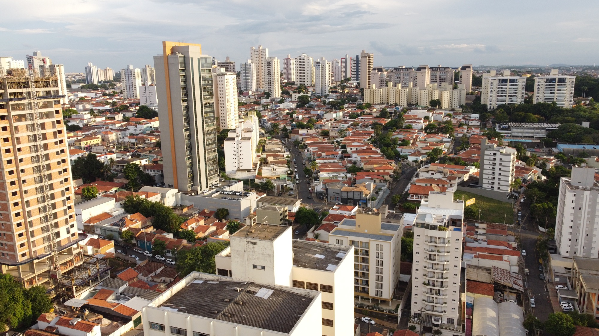 A imagem aérea mostra uma região urbana densa de Piracicaba, com uma mescla de edifícios altos e casas térreas. Em destaque, prédios residenciais modernos e construções em andamento. A paisagem revela o crescimento vertical da cidade. O céu limpo e a luz suave indicam fim de tarde.