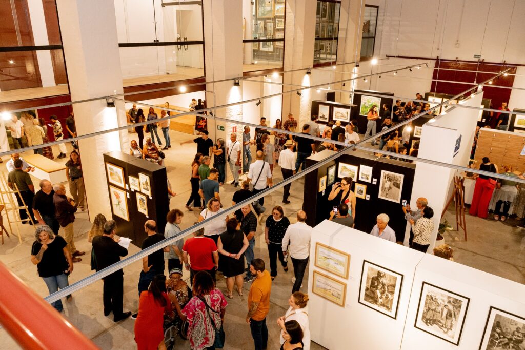 Vista aérea de uma galeria cheia de visitantes. As paredes estão cobertas por quadros e obras de arte, enquanto o espaço se organiza em divisórias. Pessoas circulam, conversam e observam os trabalhos expostos.