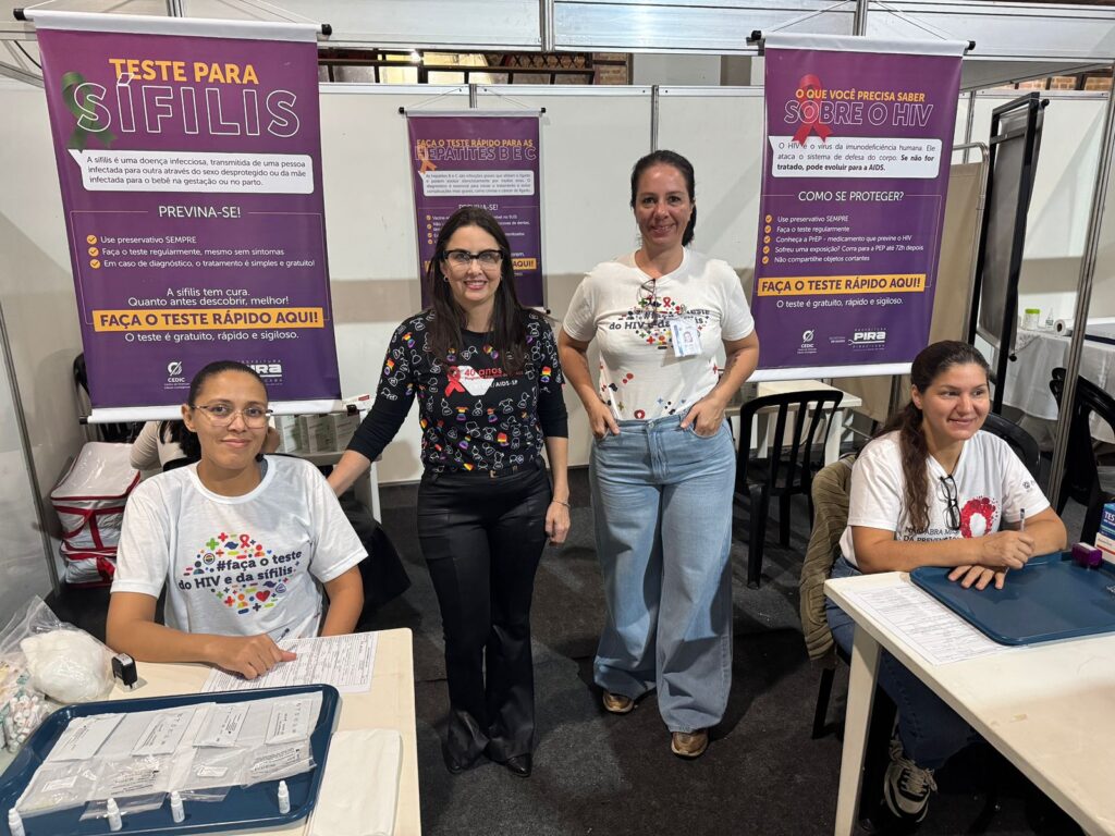 A imagem mostra um estande de conscientização sobre saúde, onde quatro pessoas estão presentes—três em pé e uma sentada à mesa. O espaço conta com dois banners grandes com informações sobre sífilis e HIV. O banner da esquerda, intitulado “TESTE PARA SÍFILIS”, destaca medidas de prevenção, como o uso de preservativos e a importância de realizar testes regularmente. Já o banner da direita, “O QUE VOCÊ PRECISA SABER SOBRE O HIV”, aborda formas de proteção, incluindo a testagem frequente e o uso de preservativos. Ambos incentivam as pessoas a realizarem testes rápidos, que são gratuitos, sigilosos e rápidos. As pessoas na imagem estão vestindo camisetas com mensagens relacionadas à saúde, e há documentos e materiais de testagem sobre a mesa. Essa cena reforça iniciativas de conscientização e promoção da saúde pública, encorajando a prevenção de infecções sexualmente transmissíveis.