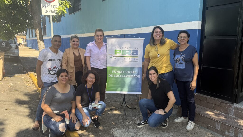A imagem mostra um grupo de pessoas em pé na frente de um letreiro que diz "Prefeitura Pira Piracicaba - Fundo Social de Solidariedade". Há sete mulheres e três homens. Estão todos sorrindo para a câmera. As mulheres estão usando uma variedade de roupas, incluindo blusas, jaquetas e calças. Os homens estão vestindo camisetas e calças. O fundo é uma parede azul e branca com uma placa que diz "Veículos Oficiais". Há uma árvore à esquerda da imagem.