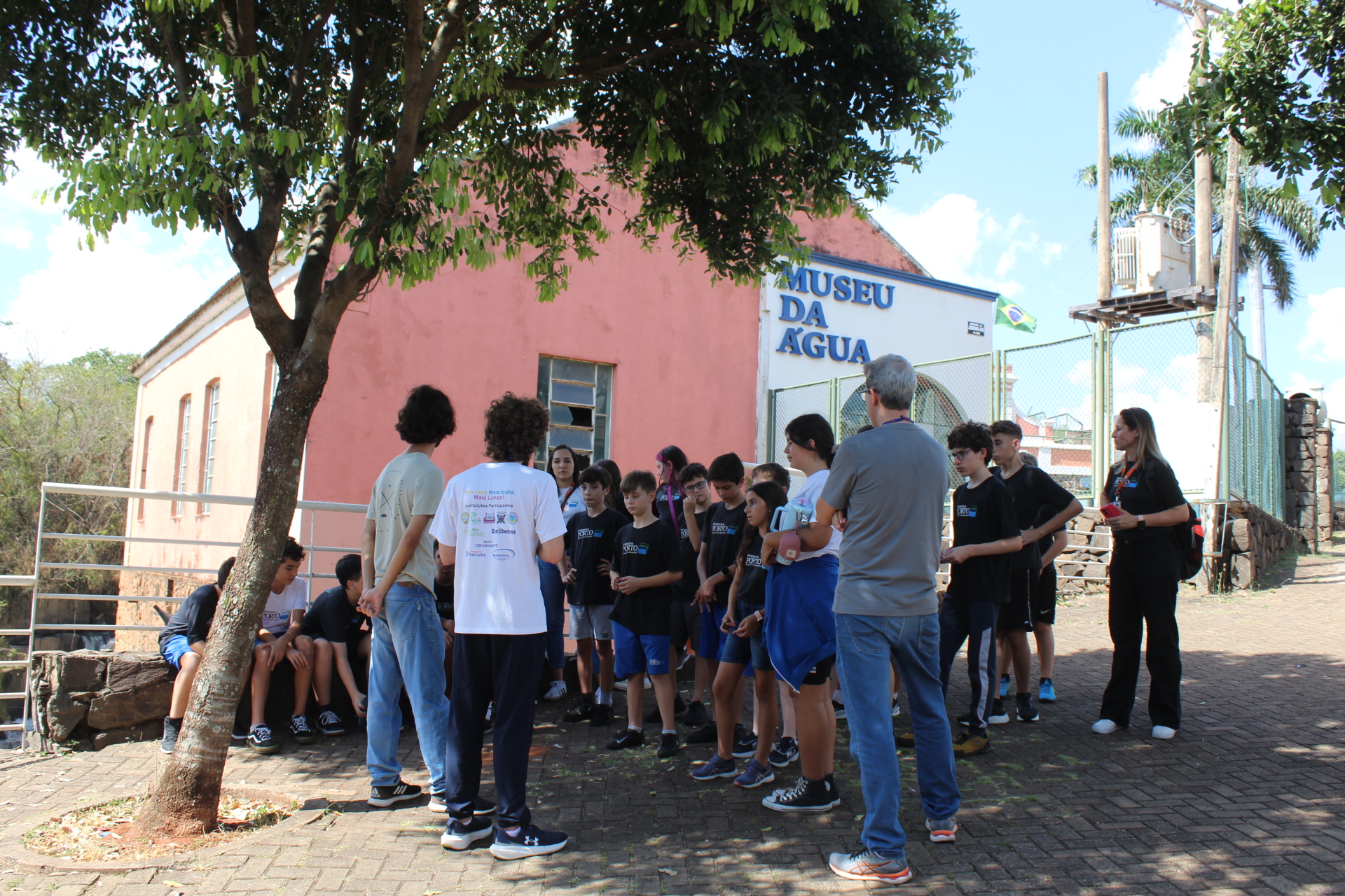 A imagem mostra um grupo de estudantes, aparentemente em uma excursão escolar, reunidos em frente ao Museu da Água. Há um adulto, possivelmente um professor ou guia, interagindo com os alunos. O grupo parece atento e interessado no que está sendo apresentado. Detalhes observáveis: * *Localização:* O Museu da Água é claramente visível na imagem, indicando o local da visita. A arquitetura do museu sugere um prédio histórico ou adaptado para o propósito do museu. Há uma cerca e uma subestação elétrica visíveis no fundo, dando contexto ao ambiente. A presença de uma árvore frondosa sugere um clima ameno e possivelmente um ambiente mais rural ou suburbano. * *Participantes:* Um grande número de adolescentes, possivelmente do ensino fundamental ou médio, estão presentes. Eles estão vestidos com roupas casuais, algumas com uniformes ou camisetas com logotipos, sugerindo uma possível organização ou evento específico. Há também pelo menos dois adultos presentes, um aparentemente interagindo com o grupo, e outro observando à distância. A roupa dos adultos sugere um papel de guia ou supervisor. * *Atividade:* A imagem captura um momento de pausa ou explicação. Os alunos estão agrupados, sugerindo atenção a alguma informação ou instrução sendo passada pelo adulto. A postura deles indica engajamento e expectativa. * *Atmosfera:* A atmosfera geral parece calma e organizada. A luz do sol indica um dia ensolarado, e a sombra da árvore fornece um local de descanso para o grupo. O ambiente parece ser seguro e acolhedor. Em resumo, a imagem retrata uma excursão escolar educativa no Museu da Água, com um grupo de alunos e adultos envolvidos em uma atividade de aprendizado informal.
