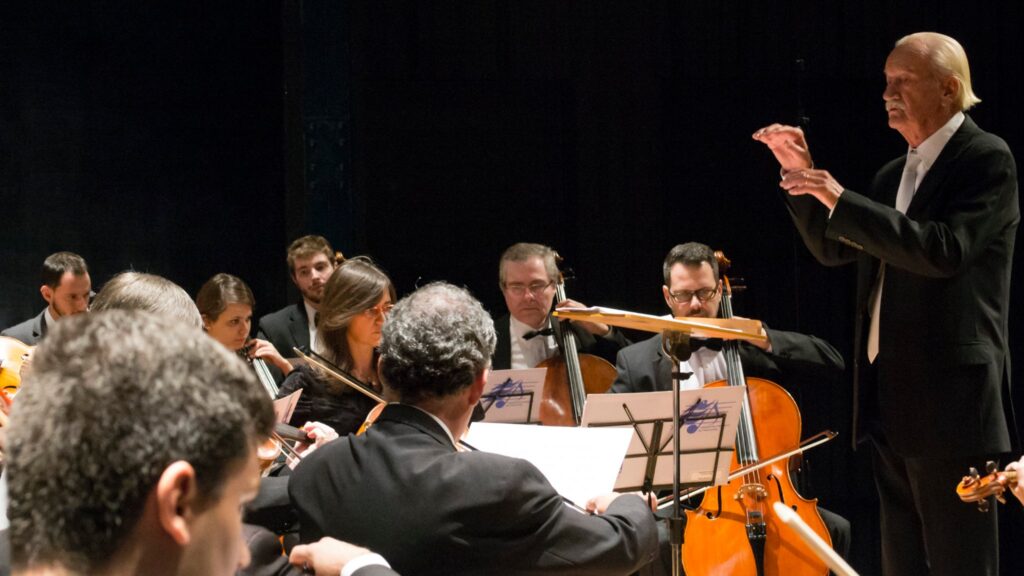 Maestro idoso, de terno preto e cabelo branco penteado para trás, está em pé regendo uma orquestra composta por músicos atentos, em trajes formais, com instrumentos de corda. A cena ocorre em palco escuro, iluminado apenas pela luz direcionada aos músicos.