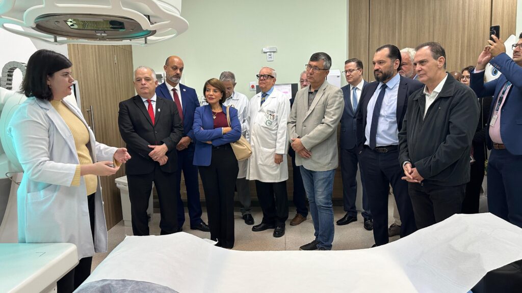 A imagem mostra um grupo de pessoas, aparentemente profissionais de saúde e autoridades, reunidos em uma sala que parece ser um novo departamento de radioterapia ou um centro de imagem médica. Uma mulher de jaleco branco está no centro da imagem, possivelmente apresentando ou explicando algo relacionado ao equipamento visível ao fundo, que parece ser um aparelho de radioterapia ou um tomógrafo. Há vários homens de terno e algumas mulheres com roupas mais formais. A atmosfera é formal e profissional, sugerindo uma inauguração, apresentação ou visita oficial a uma nova instalação médica. O tom geral da imagem é positivo, mostrando uma sensação de orgulho e realização. A presença de vários homens em posições de liderança sugere um evento importante dentro do contexto da saúde pública ou privada. A mulher em destaque, com o jaleco branco, provavelmente tem um papel crucial na apresentação ou operação do novo equipamento. A imagem carece de contexto adicional para uma interpretação mais precisa, mas a evidência visual sugere um evento relacionado à saúde, provavelmente a apresentação ou inauguração de novo equipamento médico em um hospital ou clínica.