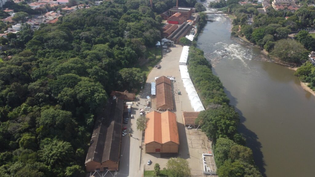 Esta imagem aérea captura uma paisagem fascinante onde um rio serpenteia por uma área densamente florestada. À esquerda do rio, há vários edifícios com telhados vermelhos, possivelmente estruturas industriais ou históricas, cercadas por árvores e vegetação. Uma área pavimentada com veículos e equipamentos também é visível, sugerindo alguma atividade humana. O rio exibe algumas corredeiras ou pequenas quedas d’água, e ao fundo, há casas residenciais, formando um equilíbrio entre natureza e desenvolvimento.