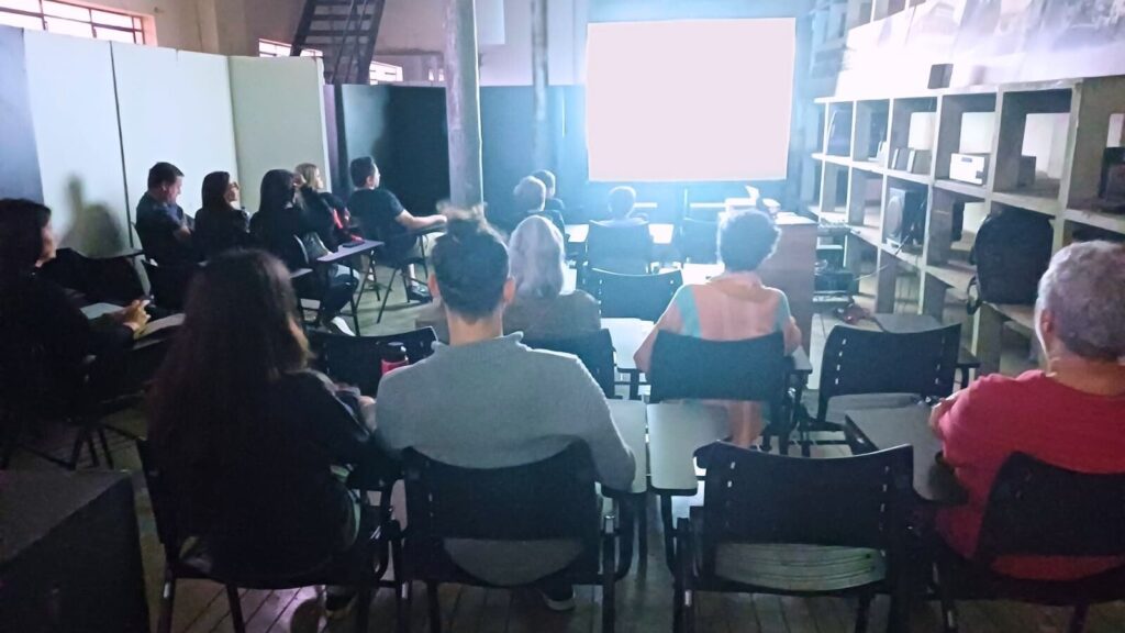 Cerca de 15 pessoas estão sentadas em uma sala escura assistindo à projeção de um filme em um telão. O ambiente é fechado, com estrutura de madeira e prateleiras ao lado.