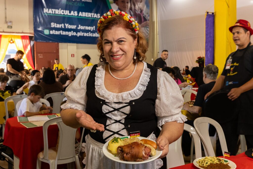 Uma voluntária caracterizada com traje típico alemão sorri enquanto apresenta um prato tradicional da culinária germânica. Na louça, destaque para o Eisbein (joelho de porco), acompanhado de salsichão, chucrute e batata. Ao fundo, o restaurante da barraca Alemanha está movimentado. A cena reflete o clima acolhedor e festivo do evento.
