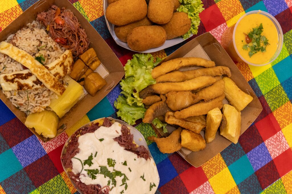 A imagem exibe uma variedade de pratos dispostos sobre uma toalha xadrez colorida, trazendo uma combinação rica e apetitosa de alimentos. Aqui estão os principais itens que aparecem: Um recipiente de papelão com arroz, carne desfiada, queijo grelhado, banana frita e fatias de abacaxi. Um prato contendo vários croquetes fritos acompanhados de folhas de alface. Outro recipiente com filés de peixe empanados e pedaços de mandioca frita, também acompanhados de alface. Uma tigela com carne desfiada coberta por um molho branco e finalizada com ervas picadas. Além disso, há um copo com um líquido amarelado, possivelmente um molho ou uma sopa, decorado com ervas. A imagem transmite uma sensação de diversidade gastronômica, provavelmente representando um tipo específico de culinária. A apresentação dos pratos destaca os diferentes sabores e texturas, tornando tudo muito convidativo.