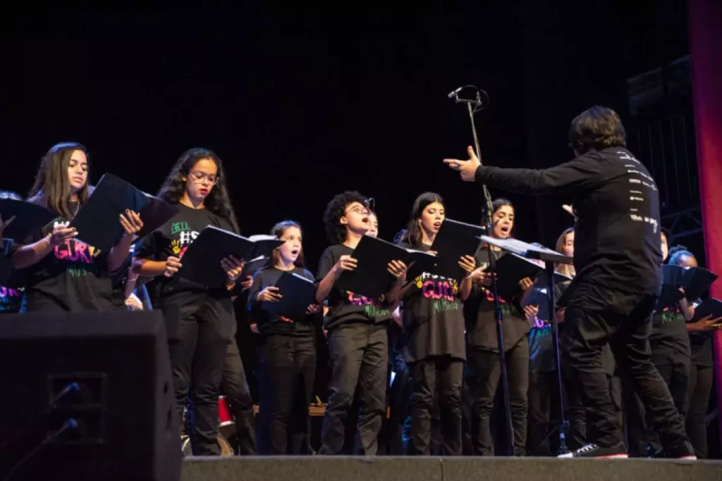 Crianças e adolescentes do projeto Guri cantam em coral sob regência de um maestro. Todos usam camisetas pretas com a logomarca do projeto e seguram partituras.
