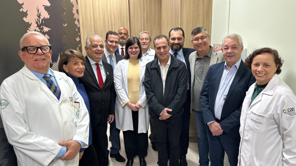 A imagem mostra um grupo de profissionais de saúde, possivelmente médicos e outros profissionais de saúde, reunidos em um corredor ou sala de espera de um hospital ou clínica. Há um total de 12 pessoas na foto, incluindo homens e mulheres de diferentes idades. A maioria está vestida com roupas formais, como ternos e jalecos brancos, indicando suas profissões. Algumas pessoas estão usando jalecos com logotipos ou identificações, sugerindo afiliação a uma instituição específica. O fundo é uma parede com um design discreto, possivelmente de madeira ou outro material similar, e um mural com um desenho de árvores de coníferas. A foto parece ter sido tirada em um ambiente profissional e formal, possivelmente para registrar um encontro ou evento relacionado à área da saúde. A expressão facial dos indivíduos é geralmente séria e profissional, embora alguns mostrem sorrisos leves. A imagem não oferece mais contexto sobre o evento ou a razão pela qual as pessoas estão reunidas.