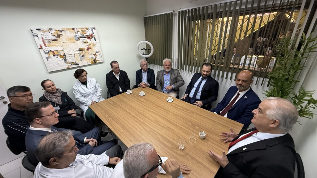 A imagem mostra uma reunião em uma sala de conferências. Há várias pessoas sentadas em torno de uma mesa de madeira retangular. A maioria das pessoas está usando roupas formais de negócios. Há uma pintura abstrata na parede atrás deles. Uma planta está em um canto da sala. A iluminação parece ser artificial, provavelmente fluorescente. A sala parece ser um escritório ou sala de reunião em um ambiente profissional. A imagem sugere uma reunião formal, possivelmente uma reunião de negócios ou uma reunião de equipe. Não há elementos que indiquem especificamente o assunto da reunião.