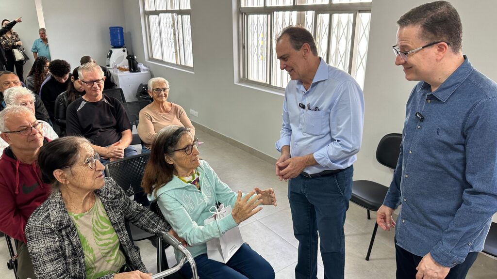 A imagem mostra uma cena em um ambiente que parece ser uma sala de espera ou um auditório. Há um grupo de idosos sentados em cadeiras, alguns com auxílio de bengalas ou cadeiras de rodas. Duas pessoas, aparentemente mais jovens, estão em pé e interagindo com os idosos sentados. Uma delas veste uma camisa azul clara e parece estar falando ou explicando algo para as pessoas mais velhas. A outra pessoa, usando uma camisa azul escura, observa a interação. Há outras pessoas no fundo, algumas em pé e outras sentadas, que parecem estar observando ou participando da reunião. O ambiente é claro e organizado, com janelas grandes que permitem a entrada de luz natural. A cena sugere uma reunião, uma visita, ou um evento comunitário voltado para a população idosa. A atmosfera parece ser calma e respeitosa.