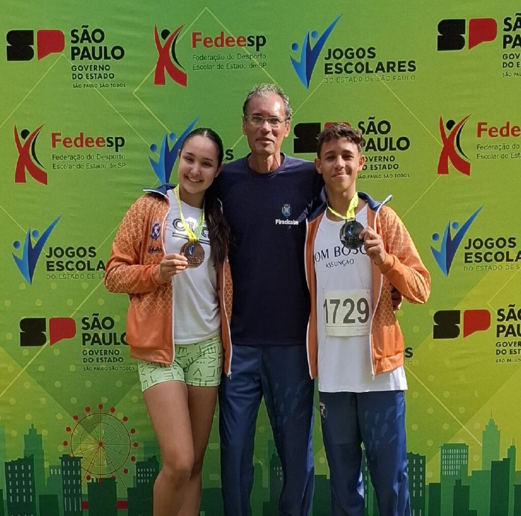 A imagem mostra três jovens atletas em frente a um painel com o logotipo dos "Jogos Escolares do Estado de São Paulo" e o logo da "Fedeesp" (Federação do Desporto Escolar do Estado de São Paulo).  Uma jovem e um rapaz estão usando jaquetas laranja e segurando medalhas de bronze e prata, respectivamente. Um homem mais velho, provavelmente um treinador ou oficial, está posicionado entre eles. Todos estão sorrindo para a câmera. O fundo apresenta um design gráfico com a silhueta de edifícios e uma roda gigante, sugerindo um contexto urbano.  Os números de identificação dos atletas são visíveis nas roupas.