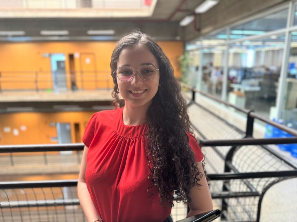 A imagem mostra uma jovem de cabelos cacheados escuros e óculos de aro fino, sorrindo para a câmera. Ela está usando uma blusa vermelha de mangas curtas e parece estar em um ambiente interno, possivelmente um prédio de escritórios ou universidade. O fundo está levemente desfocado, mas mostra uma estrutura arquitetônica moderna com corredores e janelas de vidro, sugerindo um espaço amplo e luminoso. A jovem está posicionada em um corredor com corrimão de metal, e há um leve brilho na imagem, indicando que a fotografia foi tirada em condições de boa iluminação. A imagem transmite uma sensação de profissionalismo e positividade.