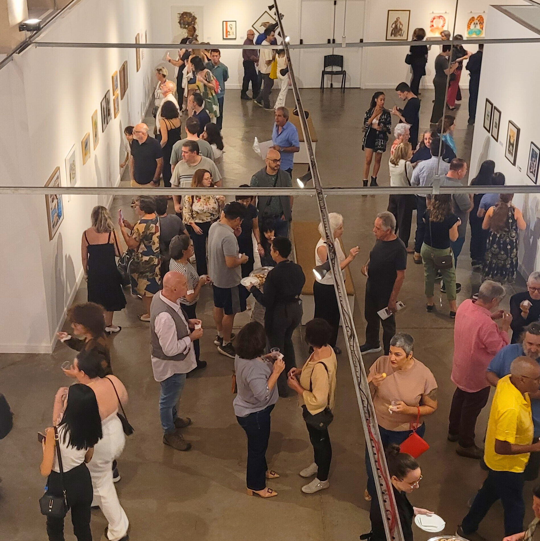 Grande galeria de arte com paredes brancas e iluminação direcionada. O espaço está lotado de visitantes que observam obras expostas nas paredes e em painéis centrais. O ambiente é vibrante e acolhedor.