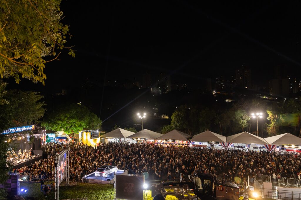 A imagem mostra um grande evento ao ar livre acontecendo à noite. À esquerda, há um palco com artistas e luzes brilhantes, e uma multidão de pessoas reunidas em frente ao palco. No lado direito, várias tendas brancas estão montadas, possivelmente servindo comida ou vendendo produtos. Ao fundo, vê-se árvores e uma paisagem urbana com prédios iluminados. A imagem transmite a atmosfera vibrante de um evento noturno com muitos participantes.
