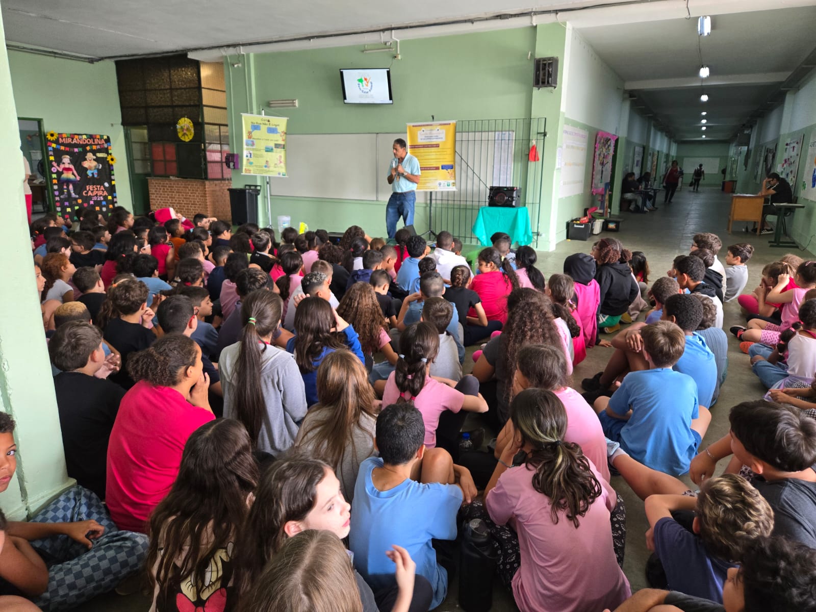 A imagem mostra uma apresentação para um grande grupo de crianças em uma escola. Um homem está no palco, falando ao microfone. As crianças estão sentadas no chão, prestando atenção. Há cartazes nas paredes e uma tela de TV. O ambiente parece ser uma escola ou um centro comunitário. A imagem sugere uma palestra ou apresentação educacional para as crianças. A atmosfera parece calma e atenta. A maioria das crianças parece estar engajada com o palestrante. Há também alguns adultos observando na multidão. A imagem transmite uma sensação de educação e aprendizagem em um ambiente escolar.