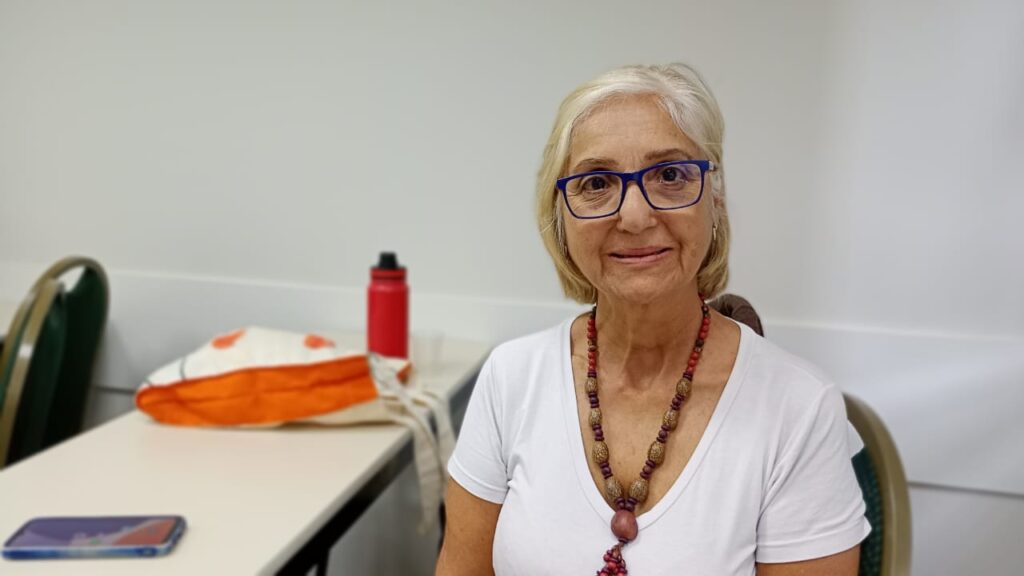 A imagem mostra uma mulher de meia-idade de cabelos grisalhos, usando óculos azuis e um colar de contas. Ela está vestindo uma camiseta branca e parece estar em uma sala de aula ou escritório, com uma mesa na frente dela com uma bolsa e um telefone celular. O fundo é uma parede branca simples. A mulher parece calma e serena, olhando diretamente para a câmera. A imagem é bem iluminada e o foco está na mulher. A qualidade da imagem é boa e os detalhes são claros. A imagem transmite uma sensação de calma e profissionalismo.