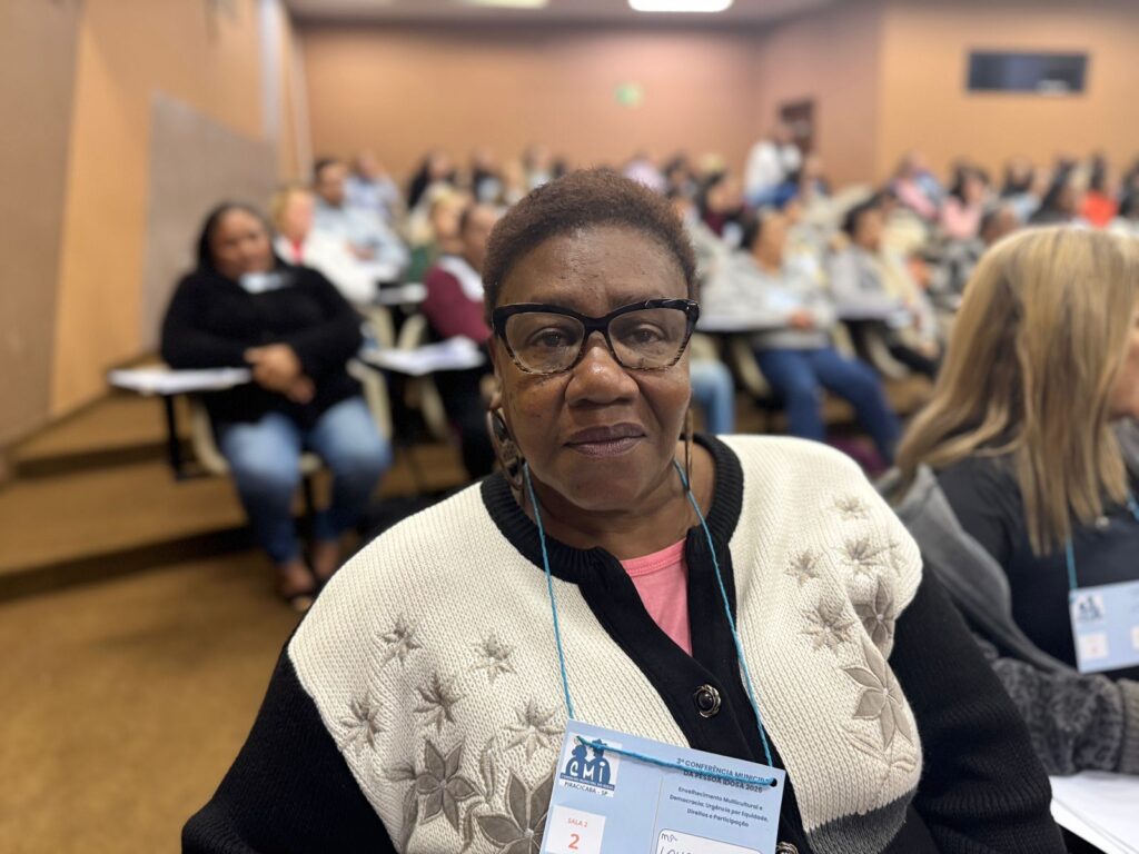 A imagem mostra um close de uma mulher negra de meia-idade sentada em uma cadeira em uma sala de aula ou auditório. Ela está usando óculos de armação escura e um suéter de tricô creme e preto com detalhes florais. Ela está usando um crachá com o texto "3ª Conferência Municipal da Pessoa Idosa 2023" visível. O fundo mostra várias outras pessoas sentadas em cadeiras, ligeiramente desfocadas, sugerindo uma reunião ou apresentação. A sala parece ser um espaço de aprendizagem formal, com paredes bege e iluminação artificial. A expressão da mulher é séria e pensativa, e ela está olhando diretamente para a câmera. A imagem é bem iluminada e o foco está na mulher em primeiro plano. A composição é simples e direta, com a mulher como o ponto focal claro da imagem.