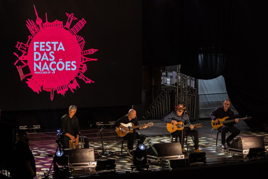 A imagem mostra uma apresentação musical ao vivo na Festa das Nações em Piracicaba, SP. No palco, há quatro músicos tocando diferentes instrumentos: dois guitarristas, um baixista e um percussionista. O palco está equipado com luzes e microfones, criando uma atmosfera vibrante. Ao fundo, uma grande tela exibe o nome do evento junto com ilustrações de monumentos famosos.