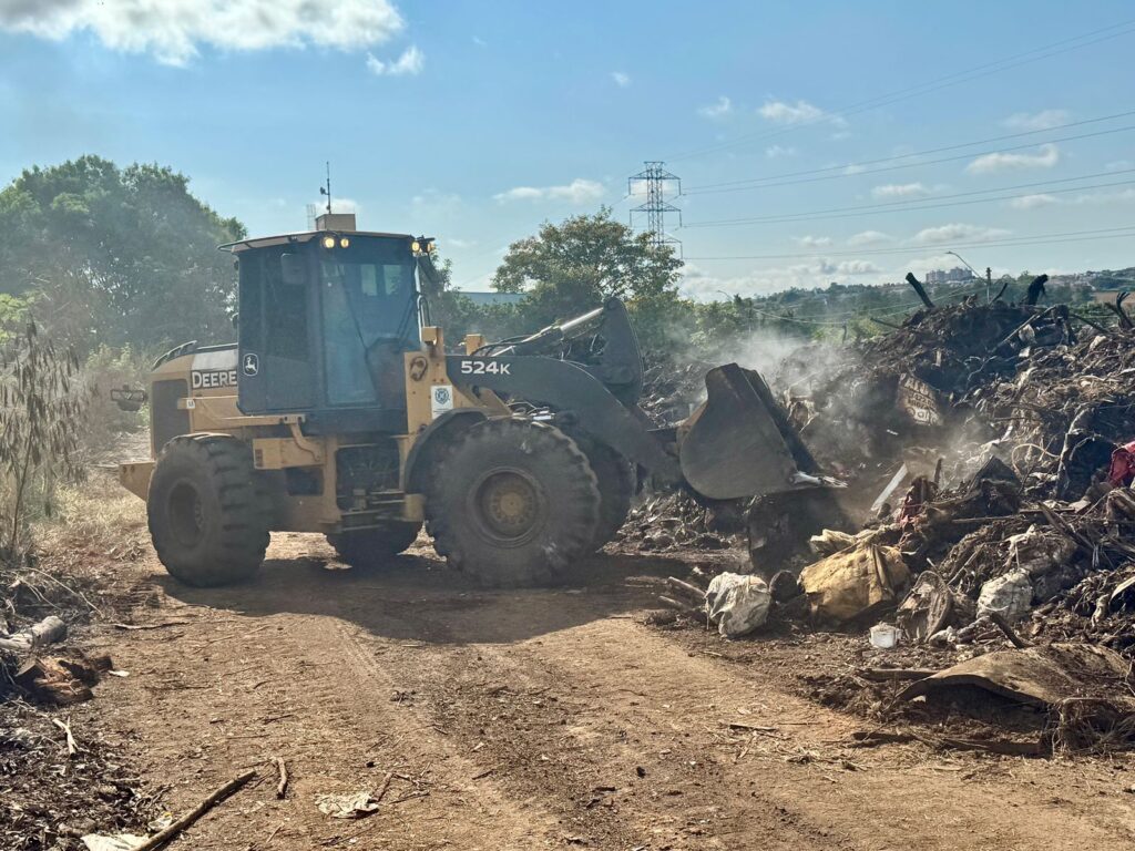 Uma pá carregadeira modelo 524K da John Deere realiza a remoção de resíduos acumulados, levantando poeira e fumaça em meio a uma grande pilha de entulho, sob céu azul com poucas nuvens.