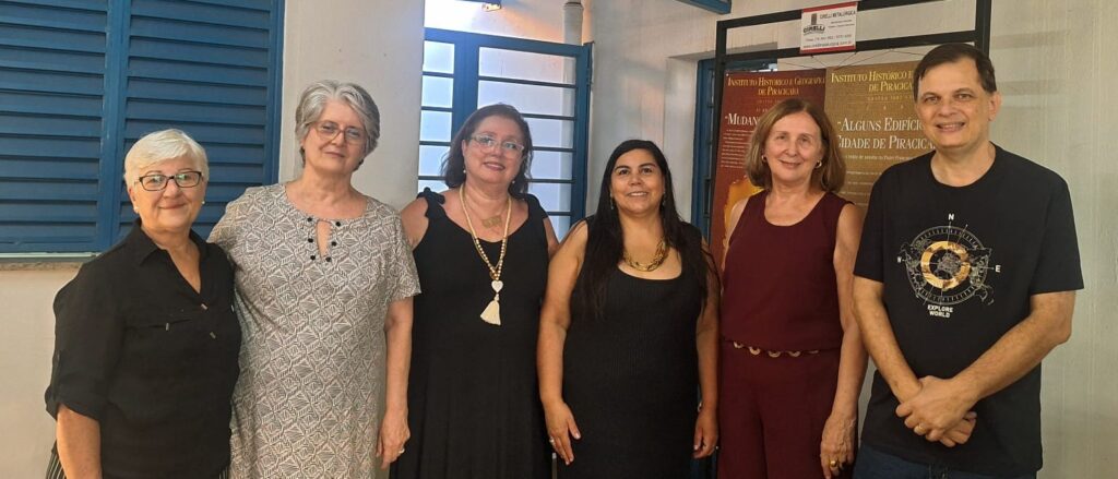 Pra Cego Ver: Seis pessoas posam sorrindo para a foto em um ambiente interno, ao lado de cartazes do Instituto Histórico e Geográfico de Piracicaba. Ao fundo, uma janela azul e portas de vidro permitem a entrada de luz. Os integrantes da imagem vestem roupas sociais casuais e demonstram alegria após o encerramento de uma eleição institucional.