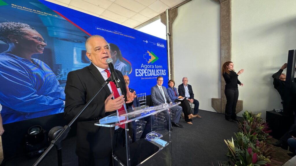 A imagem mostra uma cerimônia de inauguração ou lançamento de um projeto ou instituição relacionado ao Sistema Único de Saúde (SUS) do Brasil. Várias pessoas estão presentes, incluindo autoridades, profissionais de saúde e possivelmente representantes do governo. Podemos observar os seguintes detalhes: * *Pessoas:* Há um grupo de pessoas vestindo roupas formais, sugerindo um evento oficial. Destacam-se um homem de terno azul escuro (provavelmente uma autoridade importante) e um homem de terno escuro que parece estar discursando ou participando ativamente da cerimônia, além de uma mulher em terno azul claro e um homem de jaleco branco (provavelmente um médico). Há também outras pessoas ao fundo, algumas em trajes mais casuais. Um personagem fantasiado do SUS, com a sigla visível na roupa, também participa da cerimônia. * *Local:* O evento parece estar ocorrendo em um ambiente interno, possivelmente em um espaço amplo e moderno. Há uma parede atrás dos participantes com um banner do governo federal brasileiro, com a logo do SUS e a palavra "Brasil" em destaque. * *Ação:* O foco central da imagem é um cavalete com uma placa comemorativa, que sugere uma inauguração ou lançamento oficial. As pessoas parecem estar aplaudindo ou participando do ato inaugural. * *Contexto:* Considerando o banner com a logo do SUS e a presença de profissionais de saúde, é altamente provável que a cerimônia se relacione a algum projeto ou instituição ligada ao sistema de saúde público brasileiro. A placa no cavalete provavelmente contém informações detalhadas sobre o evento. Em resumo, a imagem captura um momento de celebração e anúncio oficial, provavelmente relacionado à expansão ou melhoria dos serviços do SUS no Brasil.