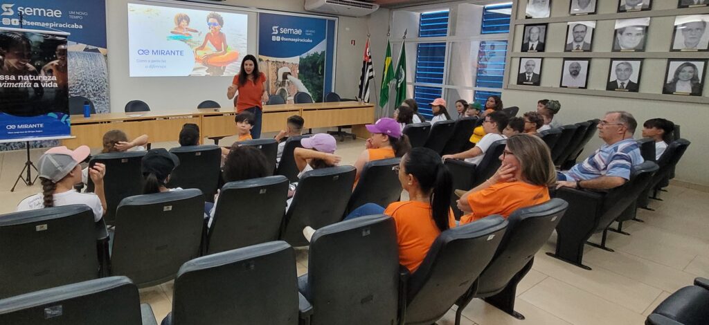 A imagem mostra uma apresentação em uma sala de palestras ou auditório. Uma mulher está apresentando um seminário ou palestra para um grupo de crianças e adolescentes, e alguns adultos. *Detalhes da imagem:* * *A apresentação:* A apresentação é sobre o "CE Mirante" e como as pessoas podem fazer a diferença. Há uma imagem de um rio ou cachoeira no slide, sugerindo possivelmente um tema ambiental ou de sustentabilidade. A apresentação é feita em português. * *A palestrante:* Uma mulher de camisa laranja está falando para a audiência, utilizando um projetor e um computador. Ela parece engajada e entusiasmada. * *A audiência:* A audiência é composta principalmente por crianças e adolescentes, sentados em cadeiras individuais em fileiras. Alguns adultos também estão presentes, incluindo alguns pais ou responsáveis. As crianças parecem atentas à apresentação. Há uma mistura de idades e gêneros na audiência. * *O local:* O local parece ser uma sala de reuniões ou auditório formal. Há uma mesa de madeira na frente, onde a palestrante está posicionada. Há também fotos emoldurando na parede, possivelmente de funcionários ou autoridades locais. Bandeiras do Brasil e possivelmente de um estado ou município também são visíveis. * *Material visual:* Há um banner ao lado da palestrante com o logotipo do "CE Mirante" e um slogan sobre a natureza. Isso reforça o tema da apresentação. *Conclusão:* A imagem sugere uma apresentação educativa ou informativa sobre o "CE Mirante", provavelmente para um público jovem, com foco em temas ambientais ou de responsabilidade social. A atmosfera parece ser formal, mas também engajadora e participativa.