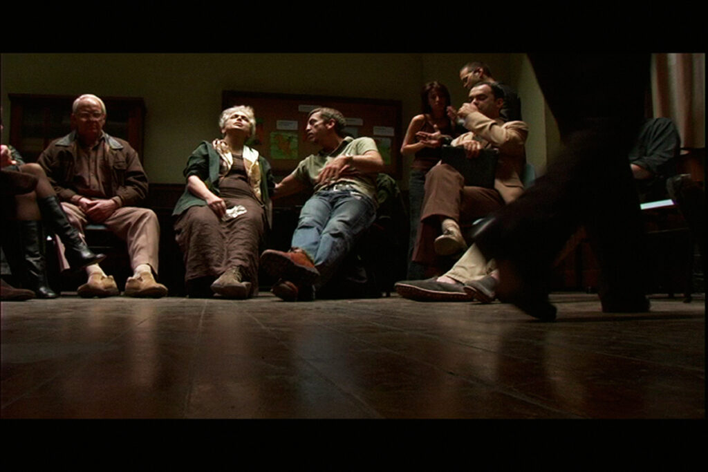 Grupo de adultos e idosos sentados em círculo participam de uma roda de conversa em um ambiente escuro. A cena é vista de baixo para cima, com foco no chão de madeira e pés dos participantes.