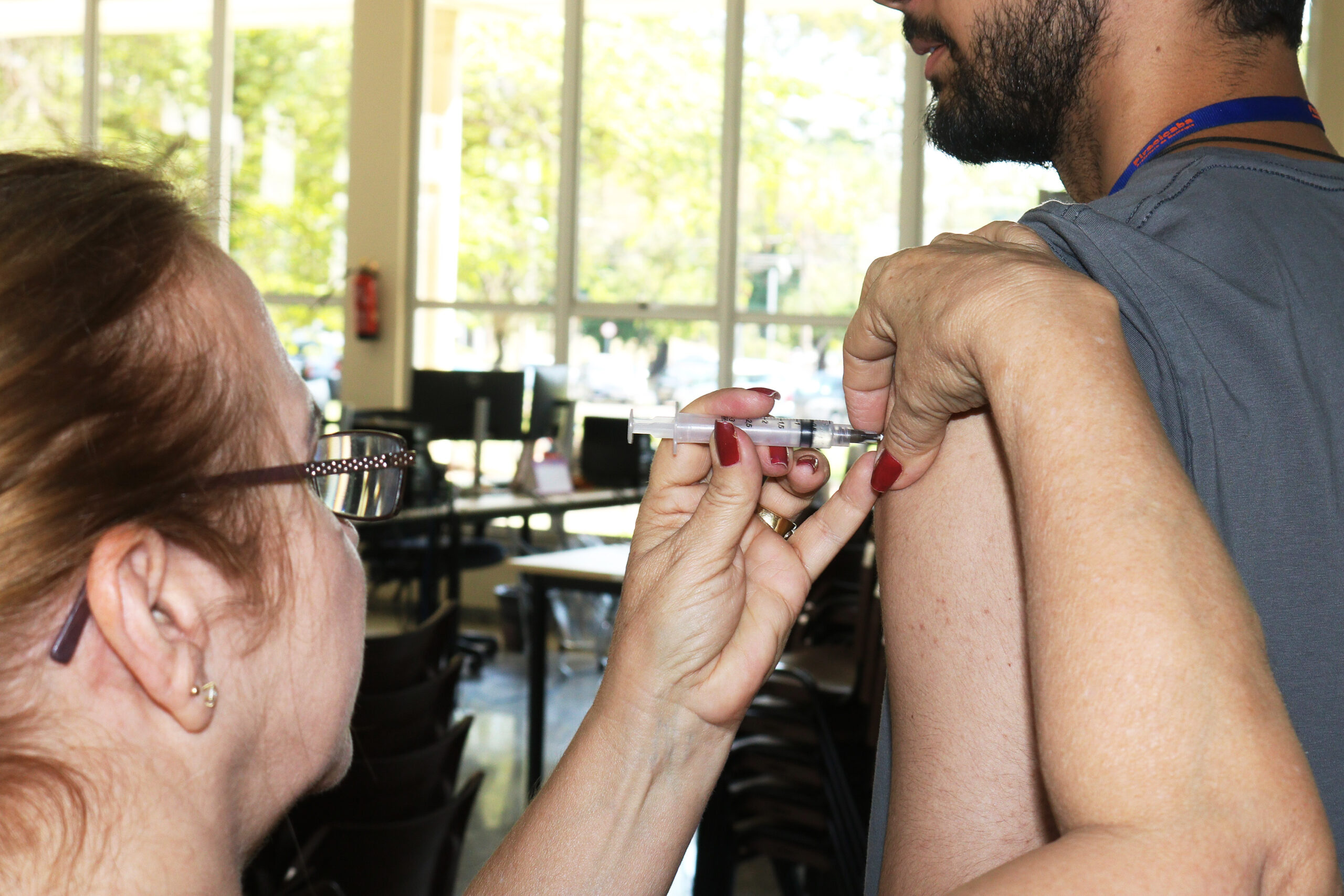 A imagem mostra uma profissional de saúde aplicando uma injeção intramuscular no braço de um paciente. A profissional, uma mulher de meia-idade com óculos, segura firmemente a seringa enquanto o paciente, um homem mais jovem, mantém o braço estendido e relaxado. O ambiente parece ser uma clínica ou local de vacinação, com computadores e cadeiras visíveis ao fundo. A janela grande ao fundo permite entrada de luz natural, indicando um ambiente claro e arejado. A imagem sugere um ato rotineiro de vacinação ou administração de medicamentos injetáveis. A atenção aos detalhes, como a postura da profissional e a expressão serena do paciente, transmitem uma sensação de profissionalismo e tranquilidade. A imagem é nítida e bem iluminada, permitindo uma observação clara do processo de injeção.