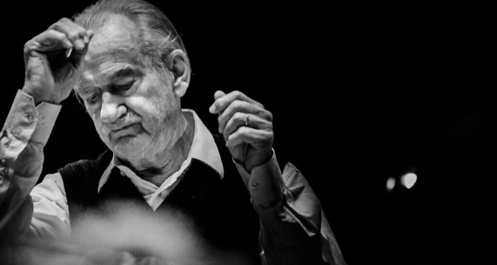 Imagem emocional do maestro Ernst Mahle com expressão concentrada, olhos semicerrados, enquanto ergue as mãos para reger. Ele veste camisa clara e colete escuro. Fundo preto, dando destaque à expressão e movimento do maestro.