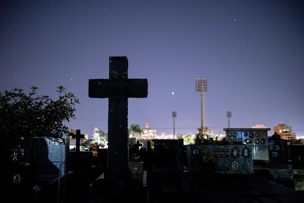 A imagem é uma fotografia noturna de um cemitério. Em primeiro plano, uma cruz de pedra escura se destaca contra um céu noturno escuro. A cruz é grande e imponente, e sua silhueta escura contrasta com o céu.Ao fundo, há uma série de lápides e túmulos, todos iluminados pela luz fraca da noite. As lápides são de vários tamanhos e formas, e algumas delas têm fotos e inscrições visíveis. No fundo, há também uma cidade iluminada, com luzes brilhantes visíveis à distância.O céu noturno é escuro e estrelado, com algumas estrelas visíveis. A atmosfera geral da imagem é sombria e reflexiva, criando uma sensação de paz e quietude. A composição da imagem é simples, mas eficaz, com a cruz como ponto focal principal.A imagem provavelmente foi tirada em um cemitério em uma cidade, com um estádio esportivo visível ao fundo. A iluminação sugere que a foto foi tirada à noite, talvez durante a madrugada. O contraste entre a escuridão do cemitério e as luzes da cidade ao fundo cria uma atmosfera contemplativa e um pouco melancólica. A presença da cruz, símbolo religioso de fé e esperança, adiciona uma camada de significado à imagem.