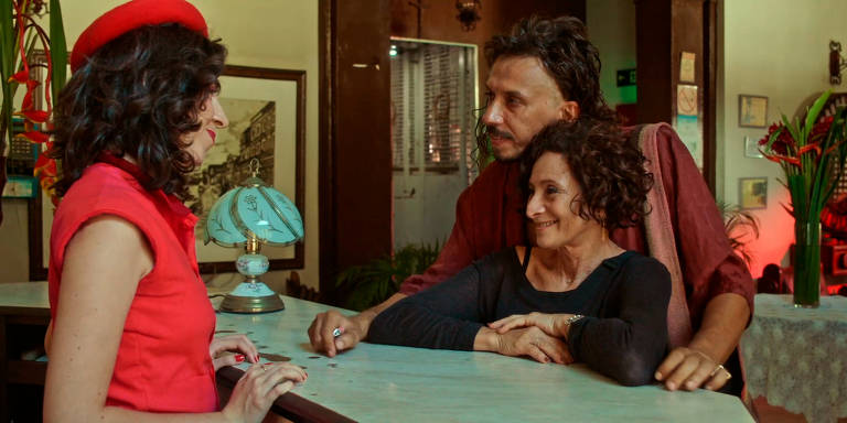 Três atores interagem no saguão de um hotel fictício. Uma mulher com boina vermelha conversa com um casal que está abraçado, evocando uma atmosfera de fantasia e drama.